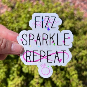 May include: A die-cut sticker with a holographic finish featuring the black text FIZZ SPARKLE REPEAT. The design includes small colorful star accents and a pink line drawing of a diamond ring at the bottom, all set against a white background.