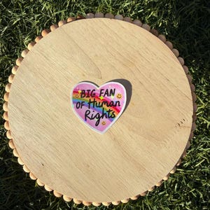 May include: A round, light-colored wooden tray with a beaded edge. A heart-shaped sticker with the text "BIG FAN OF HUMAN RIGHTS" in rainbow colors is centered on the tray. The tray is on a bed of green grass.