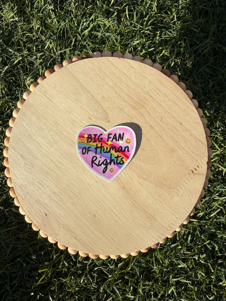 May include: A round, light-colored wooden tray with a beaded edge. A heart-shaped sticker with the text "BIG FAN OF HUMAN RIGHTS" in rainbow colors is centered on the tray. The tray is on a bed of green grass.