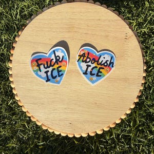 以下が含まれることがあります： 「Fuck ICE」と「Abolish ICE」というフレーズが書かれた、ハート型のステッカーが2つ表示されています。各ステッカーには虹、笑顔、青色の背景が描かれています。ステッカーは木製の丸い表面に配置されています。