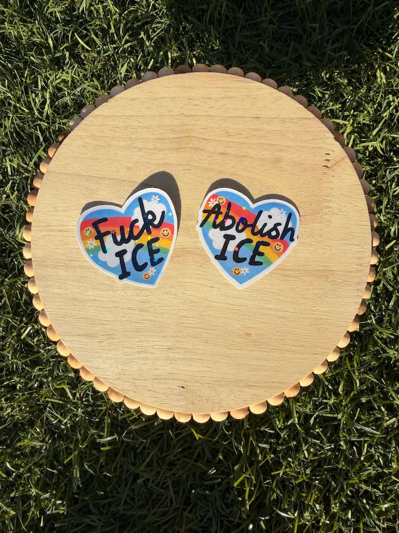 以下が含まれることがあります： 「Fuck ICE」と「Abolish ICE」というフレーズが書かれた、ハート型のステッカーが2つ表示されています。各ステッカーには虹、笑顔、青色の背景が描かれています。ステッカーは木製の丸い表面に配置されています。