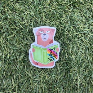 May include: A sticker featuring a cartoon dog with a grumpy expression, holding a green book. The book's cover reads "How to be an ASSHOLE and still GET TREATS" in colorful text. The sticker is set against a backdrop of green grass.