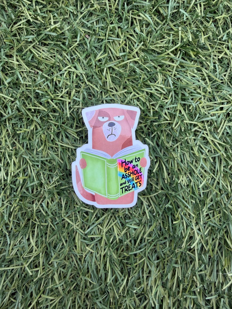 May include: A sticker featuring a cartoon dog with a grumpy expression, holding a green book. The book's cover reads "How to be an ASSHOLE and still GET TREATS" in colorful text. The sticker is set against a backdrop of green grass.