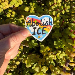 以下が含まれることがあります： 「Abolish ICE」の文字が黒、虹色、白で書かれたハート型のステッカー。ステッカーは、白い雲と小さなスマイリーフェイスのある青い空の背景を持っています。ステッカーは、緑の葉の背景の前に保持されています。