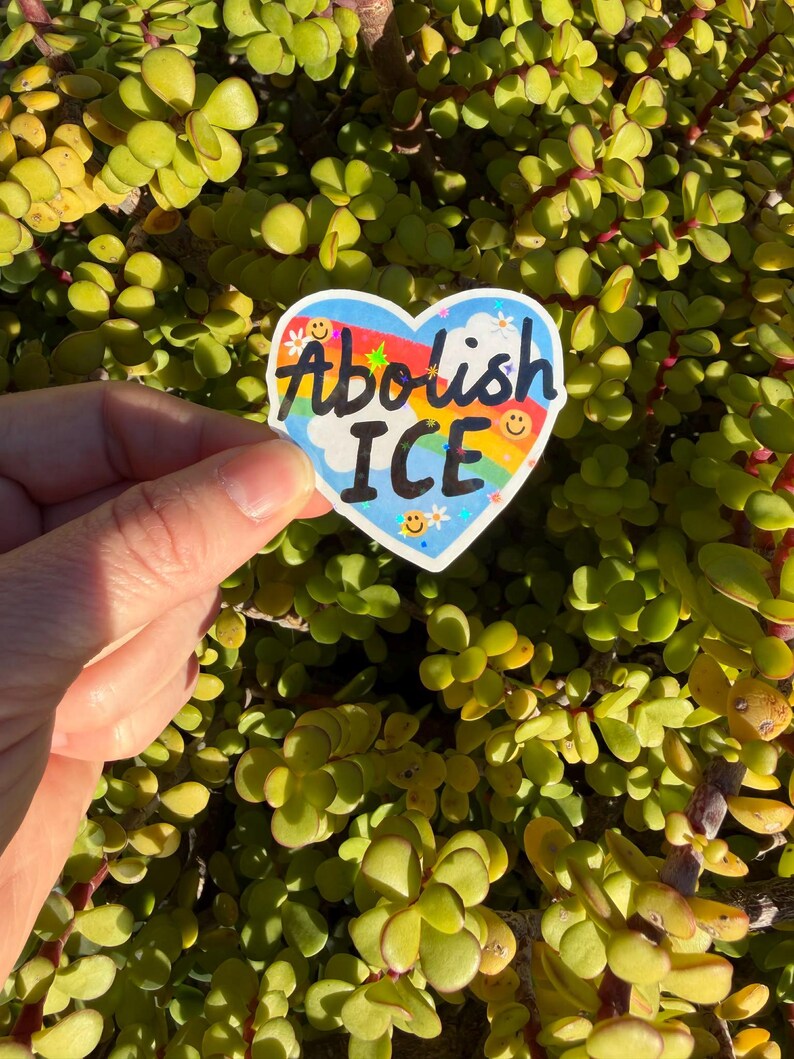 以下が含まれることがあります： 「Abolish ICE」の文字が黒、虹色、白で書かれたハート型のステッカー。ステッカーは、白い雲と小さなスマイリーフェイスのある青い空の背景を持っています。ステッカーは、緑の葉の背景の前に保持されています。