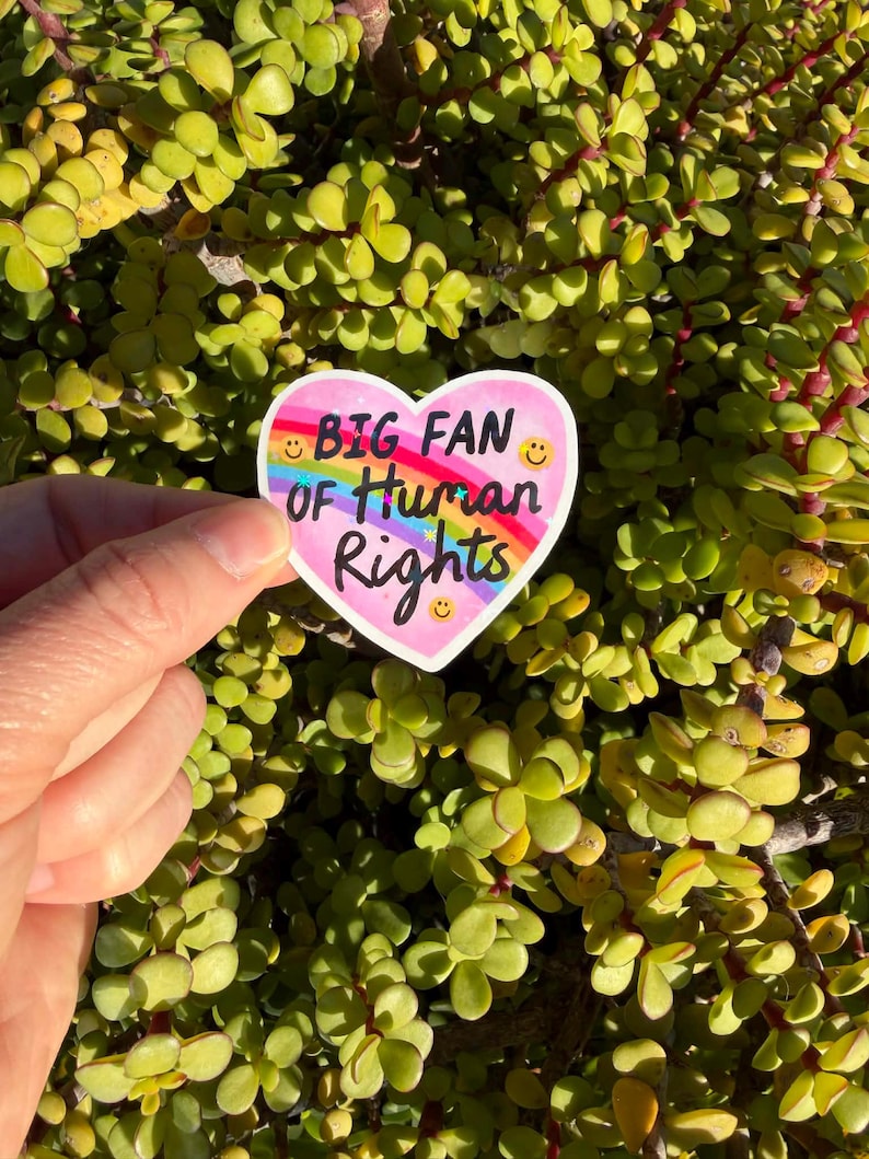May include: A heart-shaped sticker with the text "BIG FAN OF Human Rights" in black and purple. The sticker has a rainbow and smiley faces. The sticker is held in front of a background of green leaves.