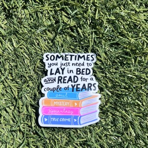 Op de afbeelding: Een sticker met een stapel boeken met de woorden "MYSTERY", "Romantasy" en "TRUE CRIME". De sticker heeft ook de tekst "SOMETIMES you just need to LAY in BED and READ for a couple of YEARS smut". De sticker staat op een groene achtergrond.