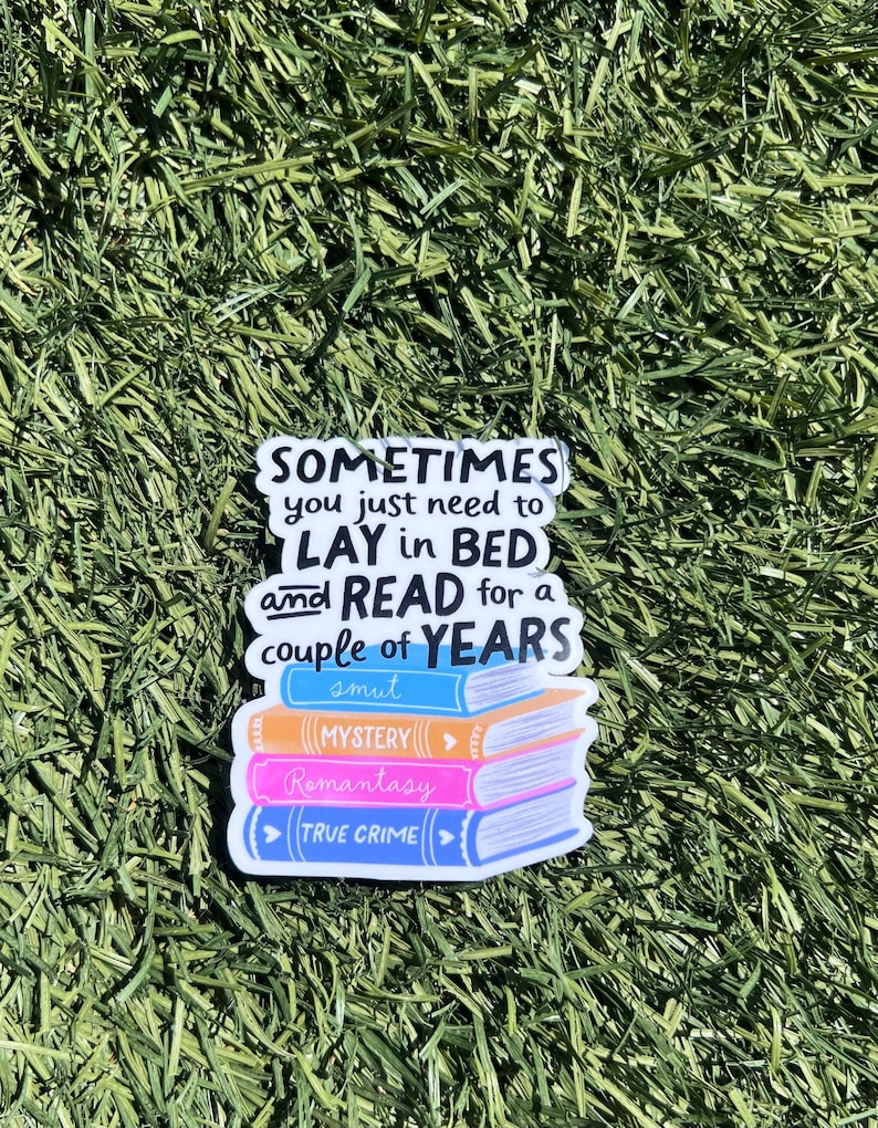 Op de afbeelding: Een sticker met een stapel boeken met de woorden "MYSTERY", "Romantasy" en "TRUE CRIME". De sticker heeft ook de tekst "SOMETIMES you just need to LAY in BED and READ for a couple of YEARS smut". De sticker staat op een groene achtergrond.
