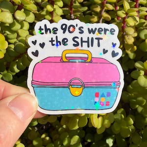 May include: A holographic sticker featuring a pink and blue plastic lunchbox with a yellow handle and latch. The text "the 90's were the SHIT" is printed in black above the lunchbox, surrounded by small black hearts and colorful star accents.