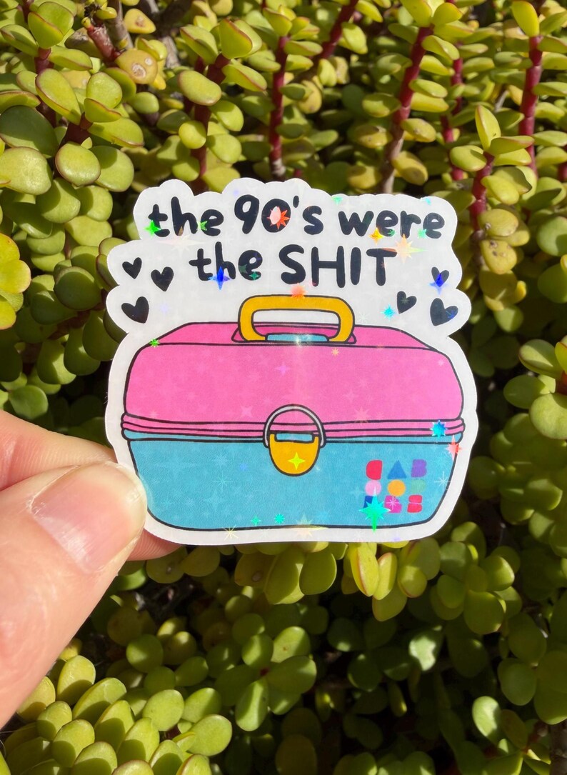 May include: A holographic sticker featuring a pink and blue plastic lunchbox with a yellow handle and latch. The text "the 90's were the SHIT" is printed in black above the lunchbox, surrounded by small black hearts and colorful star accents.