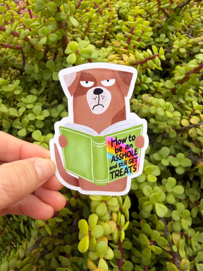 May include: A sticker featuring a cartoon dog with a grumpy expression, holding a green book. The book's title reads "How to be an ASSHOLE and still GET TREATS" in colorful text. The sticker is held against a backdrop of green foliage.