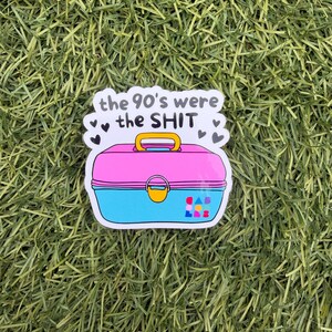 May include: A sticker featuring a pink and blue plastic lunchbox with a yellow handle and latch. The text above the lunchbox reads, the 90's were the SHIT, surrounded by small black hearts. The lower right corner features a small colorful logo.