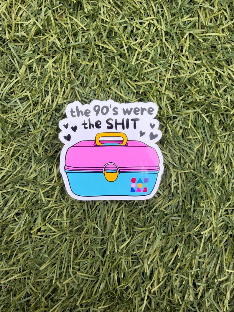 May include: A sticker featuring a pink and blue plastic lunchbox with a yellow handle and latch. The text above the lunchbox reads, the 90's were the SHIT, surrounded by small black hearts. The lower right corner features a small colorful logo.