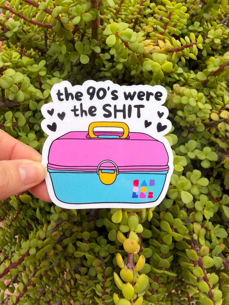 May include: A die-cut sticker featuring a pink and blue plastic lunchbox with a yellow handle and latch. The text "the 90's were the SHIT" is printed in black above the lunchbox, surrounded by small black hearts and colorful geometric shapes.