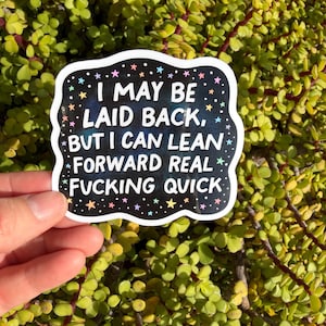 May include: A sticker with a black background and white border, featuring the text "I MAY BE LAID BACK, BUT I CAN LEAN FORWARD REAL FUCKING QUICK." The text is in white, with colorful stars and dots around the border.