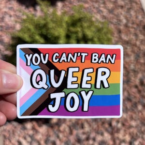 May include: A rectangular sticker with the text "YOU CAN'T BAN QUEER JOY" in white, bold letters. The sticker features the Progress Pride flag and a rainbow design. The background is blurred.