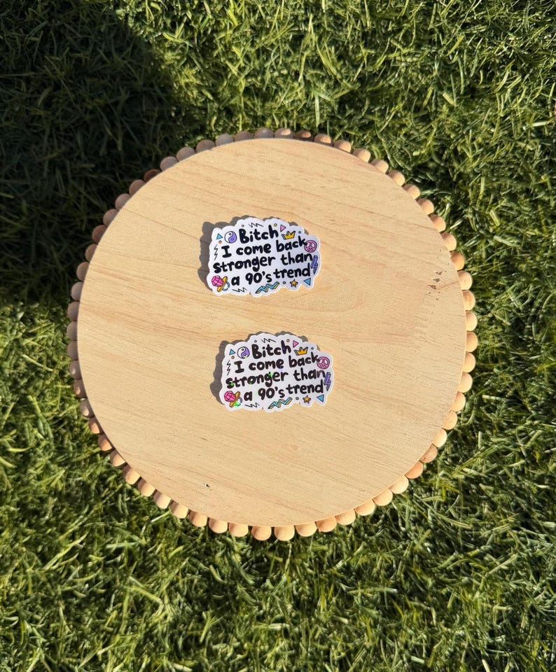 May include: Two white die-cut stickers featuring the black text "Bitch I come back stronger than a 90's trend" surrounded by colorful small graphic icons including a crown, lightning bolt, peace sign, and geometric shapes on a light wood surface.