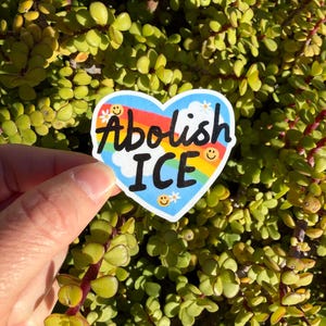 以下が含まれることがあります： 虹と青い空を背景に、黒い文字で「Abolish ICE」と書かれたハート型のステッカー。ステッカーには笑顔と花のアクセントが含まれています。ステッカーは緑の植物の背景の前に保持されています。