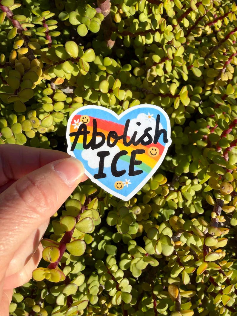 以下が含まれることがあります： 虹と青い空を背景に、黒い文字で「Abolish ICE」と書かれたハート型のステッカー。ステッカーには笑顔と花のアクセントが含まれています。ステッカーは緑の植物の背景の前に保持されています。