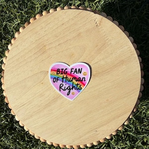 May include: A round wooden tray with a decorative beaded edge. A heart-shaped sticker with the text "BIG FAN OF HUMAN RIGHTS" and a rainbow design is centered on the tray. The tray is on a bed of green grass.