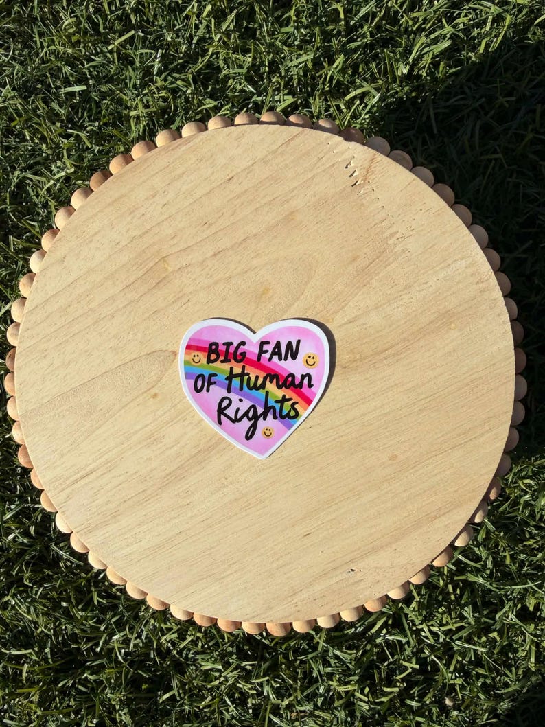 May include: A round wooden tray with a decorative beaded edge. A heart-shaped sticker with the text "BIG FAN OF HUMAN RIGHTS" and a rainbow design is centered on the tray. The tray is on a bed of green grass.