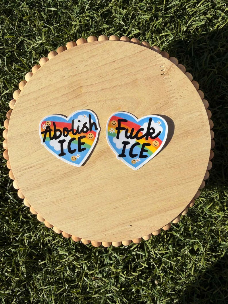以下が含まれることがあります： 「Abolish ICE」と「Fuck ICE」というフレーズが黒い文字で書かれた、ハート型のステッカーが2つ。各ステッカーは、白い雲、虹、笑顔の花が描かれた青い空の背景を持っています。ステッカーは木製のトレイの上にあります。