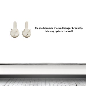 May include: Two white wall hanger brackets with four small pins each. The brackets are designed to be hammered into a wall. Text on the image reads: "Please hammer the wall hanger brackets this way up into the wall."
