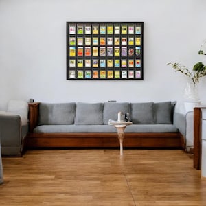 Op de afbeelding: Een ingelijste display van beoordeelde ruilkaarten hangt boven een grijze bank met houten bekleding. De kaarten zitten in zwarte hoesjes, gerangschikt in een raster. Voor de bank staat een kleine, witte tafel met een decoratieve voet.