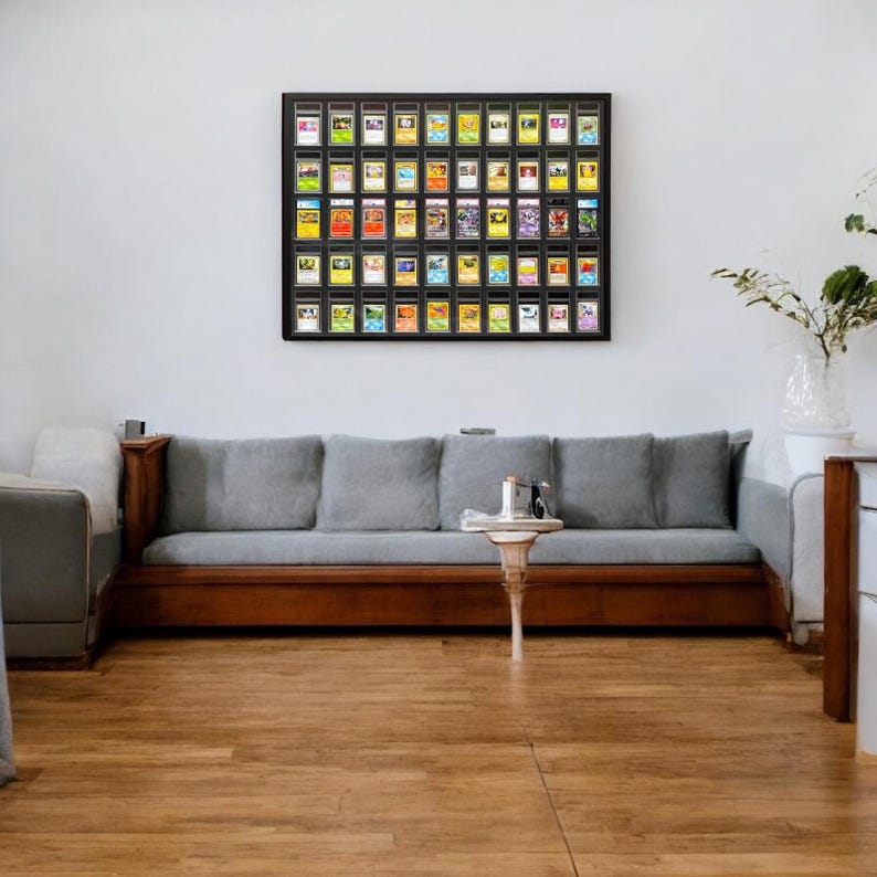 Op de afbeelding: Een ingelijste display van beoordeelde ruilkaarten hangt boven een grijze bank met houten bekleding. De kaarten zitten in zwarte hoesjes, gerangschikt in een raster. Voor de bank staat een kleine, witte tafel met een decoratieve voet.