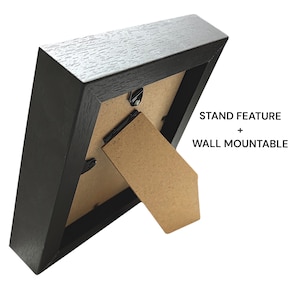 May include: A black picture frame with a stand feature and wall mountable design. The frame has a textured surface and a neutral-colored backing. The text "STAND FEATURE + WALL MOUNTABLE" is visible.