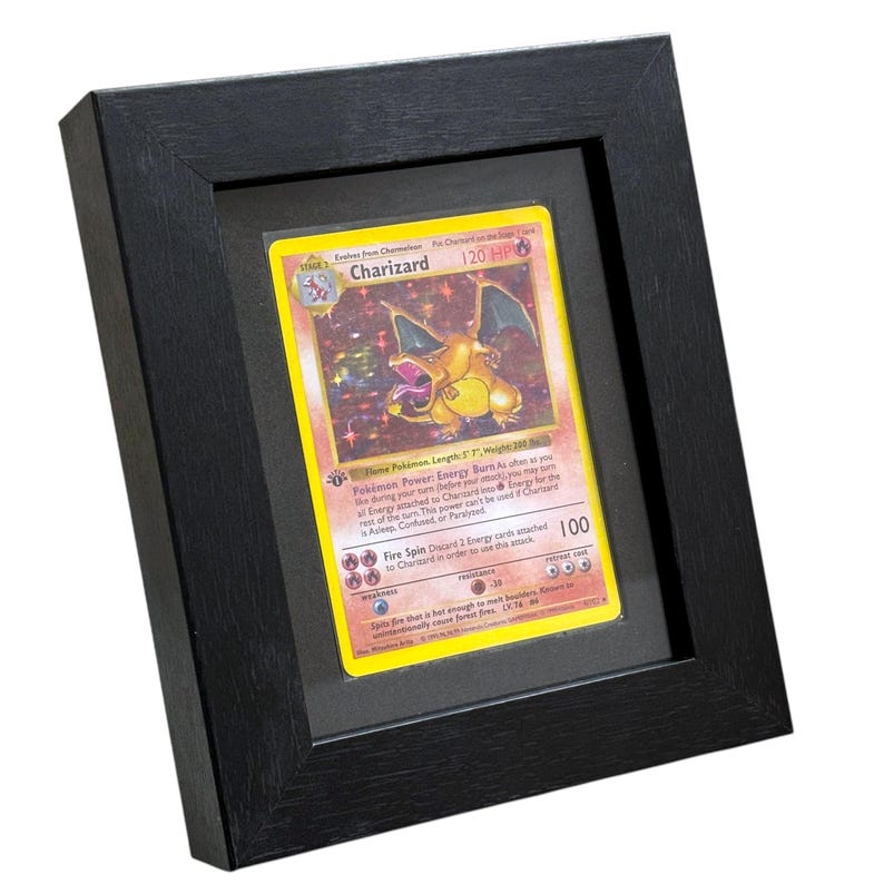 May include: A framed Charizard Pok&eacute;mon card. The card is yellow with a colorful illustration of a dragon-like creature. The black frame has a matte finish and a simple, modern design. The card is protected by a clear cover.