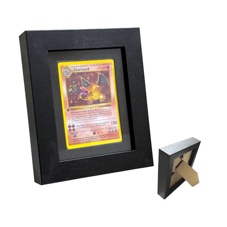 May include: A black framed display case holding a Charizard Pokemon card. The card is yellow with a Charizard illustration and text. A smaller, identical frame is next to the larger one. The frames are made of wood.