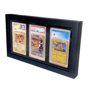 Op de afbeelding: Een zwart omkaderde display met drie beoordeelde Pok&eacute;mon-ruilkaarten. De kaarten tonen Pikachu en andere personages. De behuizing is rechthoekig en heeft een matte afwerking, geschikt voor wandmontage.