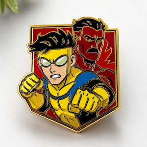 May include: A gold-rimmed enamel pin featuring a graphic illustration of a character in a yellow and blue suit with goggles in the foreground, and a red-toned character with a mustache in the background, set against a solid red shield-shaped backdrop.