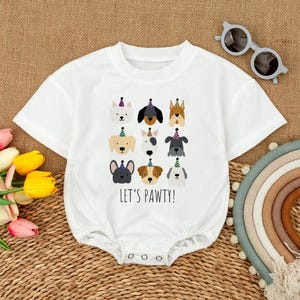 Pu&ograve; includere: Body da bambino bianco con scollo tondo e chiusura con bottoni a pressione. La parte anteriore presenta illustrazioni di varie razze di cani con cappelli da festa, con la scritta "LET'S PAWTY!" in nero. Un outfit divertente e festoso per un bambino.