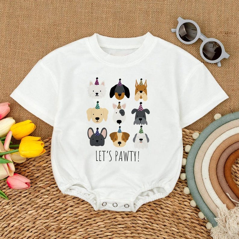 Pu&ograve; includere: Body da bambino bianco con scollo tondo e chiusura con bottoni a pressione. La parte anteriore presenta illustrazioni di varie razze di cani con cappelli da festa, con la scritta "LET'S PAWTY!" in nero. Un outfit divertente e festoso per un bambino.