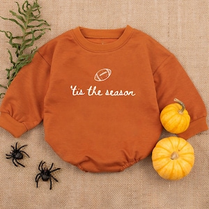 May include: An orange baby bodysuit with long sleeves and the text "'tis the season" and a football graphic. Two small pumpkins and two toy spiders are next to the bodysuit on a burlap surface.
