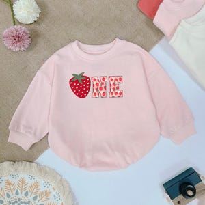 May include: Pink long-sleeved sweatshirt with a strawberry and the word "ONE" in strawberry print.