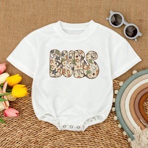 May include: White baby bodysuit with the word "BUBS" in a patterned design featuring horseshoes, cacti, and stars. The short-sleeved onesie has snap closures at the bottom and a round neckline. Sunglasses and a rainbow are in the background.