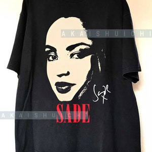 Może przedstawiać: Czarny T-shirt z białym siluetem twarzy Sade i jej podpisem w kolorze białym. Tekst "SADE" jest czerwonymi literami.