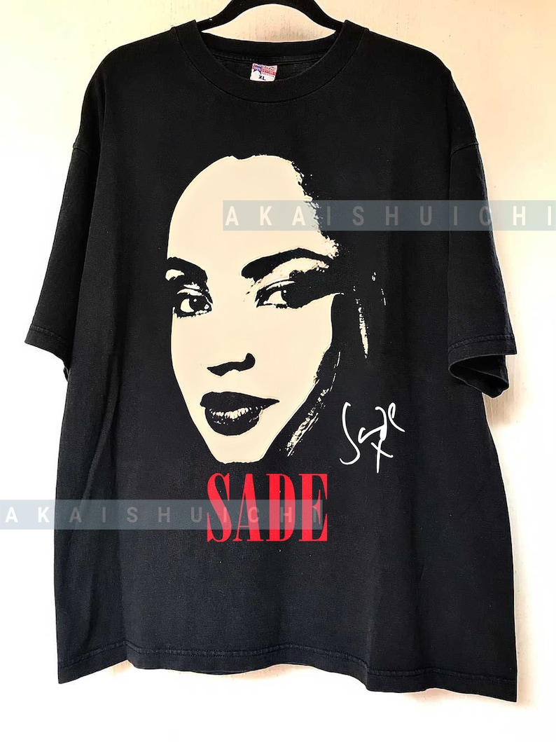 Może przedstawiać: Czarny T-shirt z białym siluetem twarzy Sade i jej podpisem w kolorze białym. Tekst "SADE" jest czerwonymi literami.