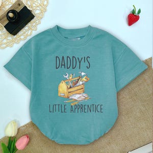 May include: A teal short-sleeved baby bodysuit with the text "DADDY'S LITTLE APPRENTICE" and a watercolor illustration of a toolbox with tools. The bodysuit is made of soft material.