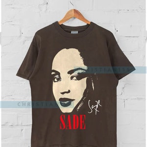 Może przedstawiać: Brązowy T-shirt z czarno-białym obrazkiem Sade. Obrazek to zbliżenie jej twarzy i zawiera jej podpis w kolorze czarnym poniżej. Słowo "SADE" jest w czerwonych literach pod podpisem.