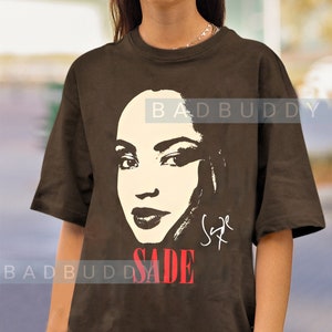 Może przedstawiać: Brązowy T-shirt z czarno-białym obrazkiem Sade z przodu. Obrazek to zbliżenie twarzy Sade, a tekst "SADE" jest nadrukowany na czerwono pod obrazkiem. Tekst "BADBUDDY" jest nadrukowany na biało na plecach koszulki.
