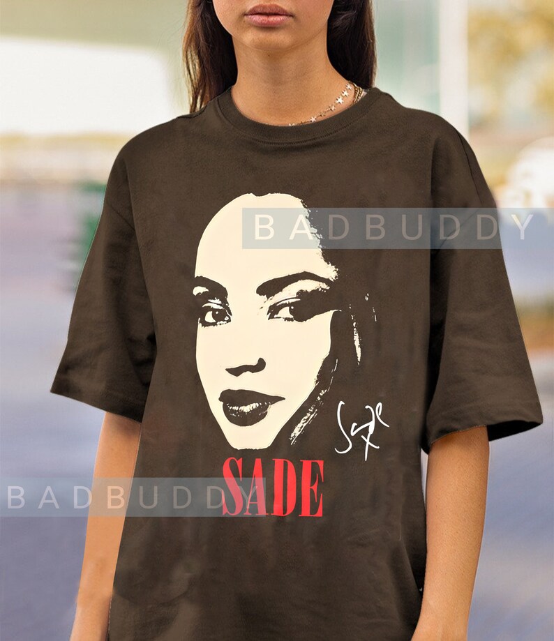 Może przedstawiać: Brązowy T-shirt z czarno-białym obrazkiem Sade z przodu. Obrazek to zbliżenie twarzy Sade, a tekst "SADE" jest nadrukowany na czerwono pod obrazkiem. Tekst "BADBUDDY" jest nadrukowany na biało na plecach koszulki.