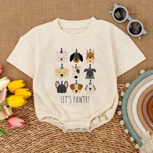 Pu&ograve; includere: Pagliaccetto per neonati color crema con girocollo e maniche corte. La parte anteriore presenta una grafica di vari volti di cani con cappelli da festa, con la scritta "LET'S PAWTY!" stampata sotto. Il pagliaccetto ha chiusure a scatto sul fondo.