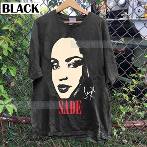 Może przedstawiać: Czarny T-shirt z czarno-białym obrazkiem Sade, piosenkarki, z jej podpisem pod obrazkiem. Tekst "SADE" jest czerwonymi literami pod podpisem.