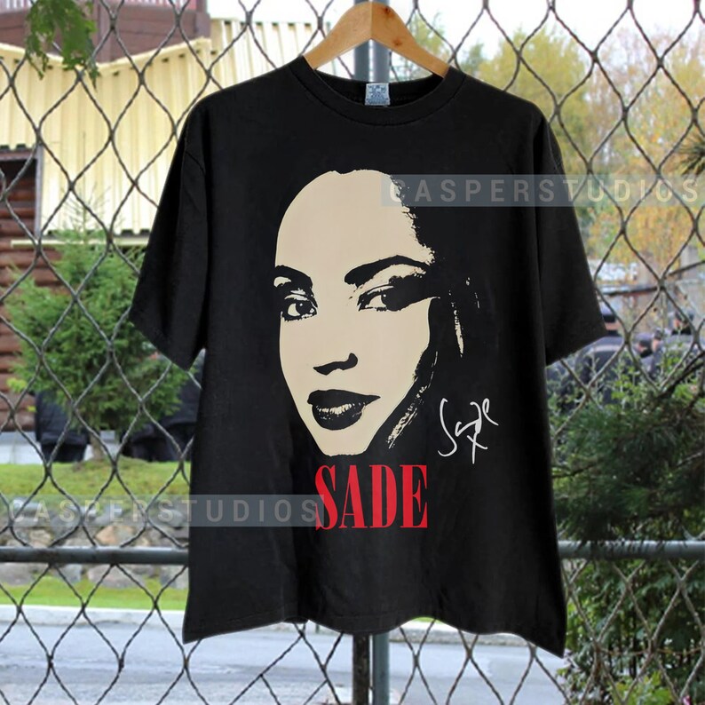 Może przedstawiać: Czarny T-shirt z czarno-białym obrazkiem Sade, piosenkarki, z jej podpisem w kolorze białym. Słowo "SADE" jest nadrukowane na czerwono pod obrazkiem.