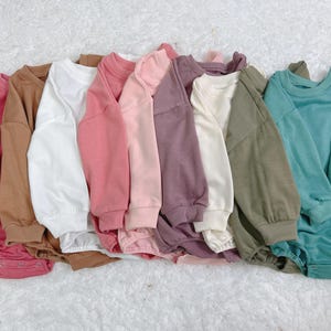 Pu&ograve; includere: Una collezione di tutine per bambini in vari colori uniti, tra cui rosa, marrone, bianco e verde acqua. Ogni tutina ha maniche lunghe e scollo tondo. Gli indumenti sono disposti su una superficie bianca strutturata.
