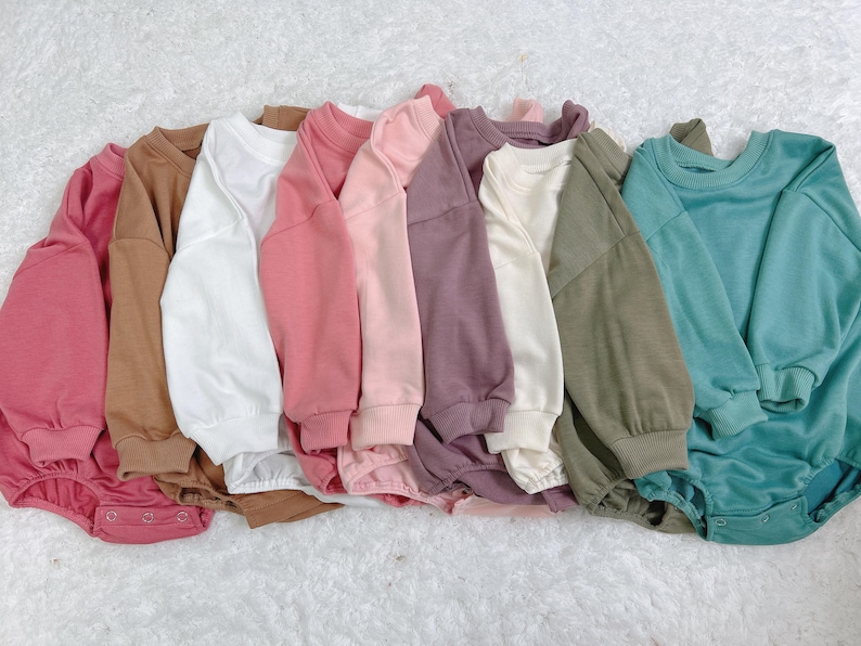 Pu&ograve; includere: Una collezione di tutine per bambini in vari colori uniti, tra cui rosa, marrone, bianco e verde acqua. Ogni tutina ha maniche lunghe e scollo tondo. Gli indumenti sono disposti su una superficie bianca strutturata.